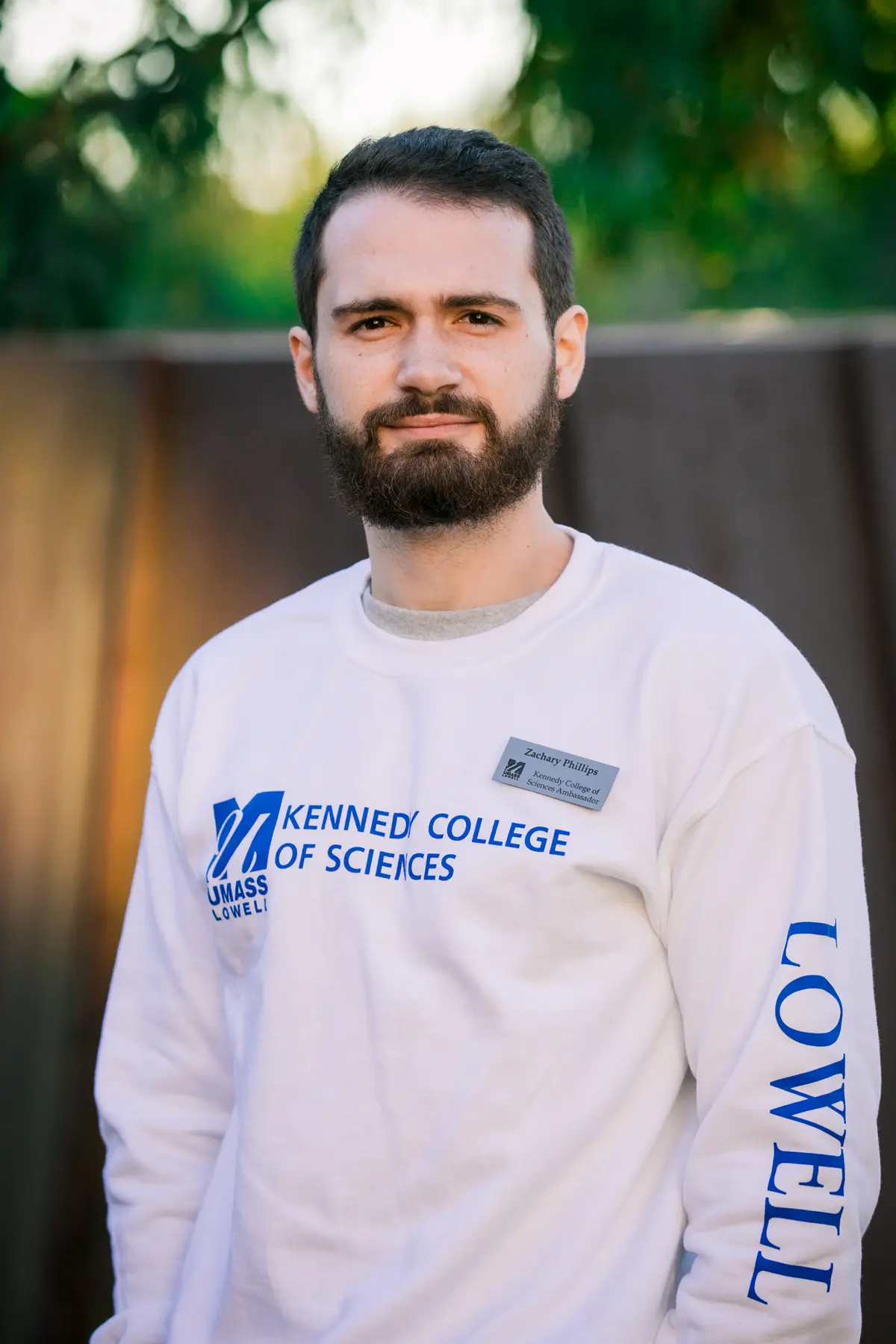 A young man with dark hair and a beard poses for a photo outside.